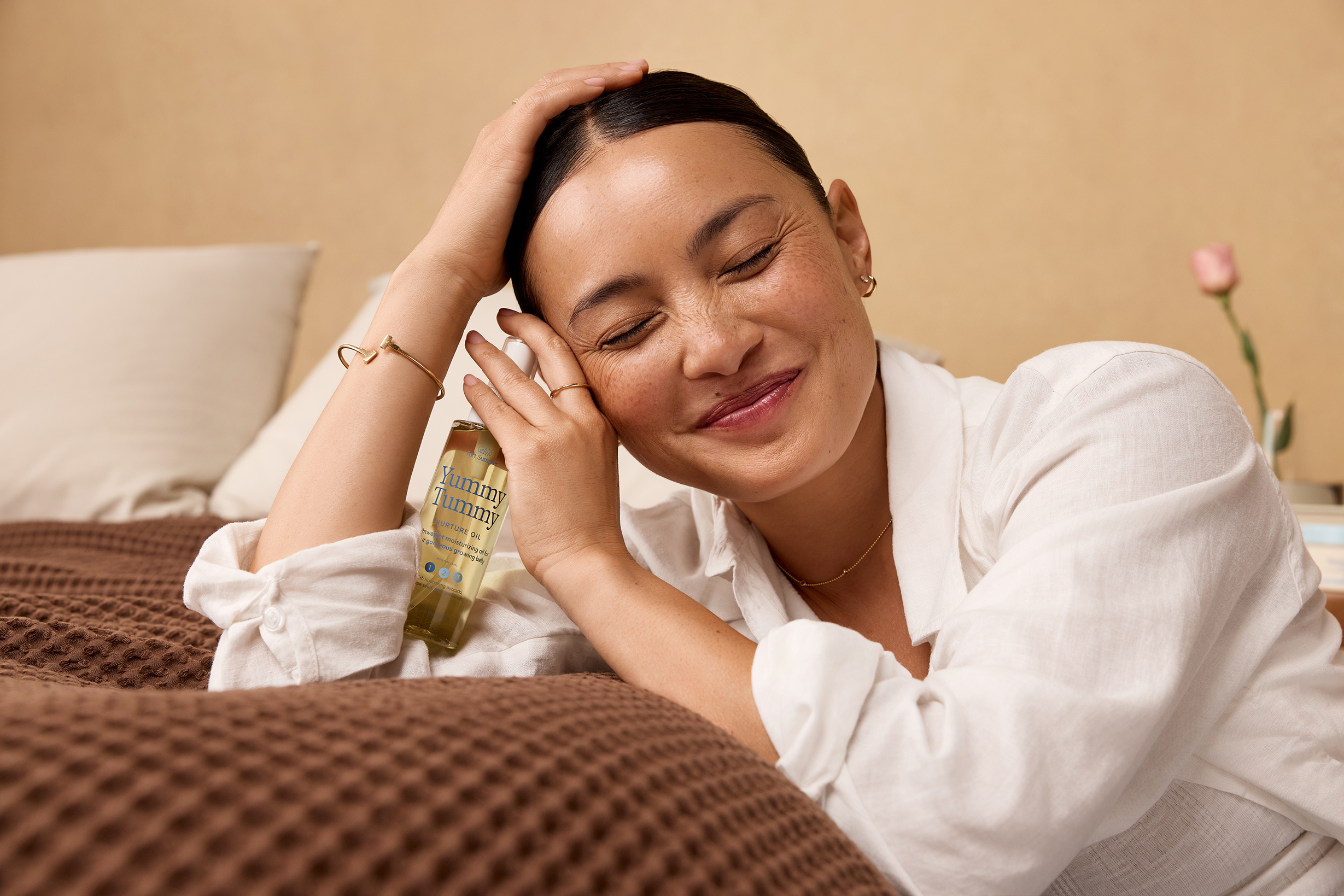 Model with her eyes closd, leaning on a bed holding the Yummy Tummy Mother Oil